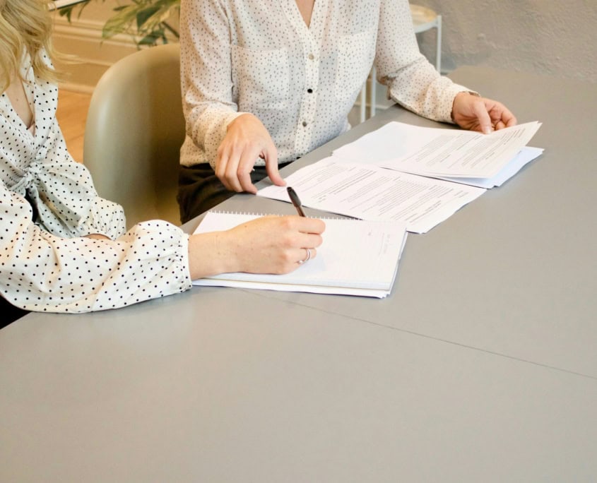 Two people in polka dot shirts review and write notes on documents at a grey table.