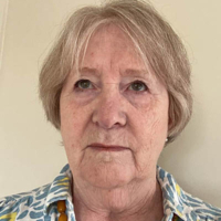 Older woman with short grey hair, wearing a patterned shirt and yellow necklace, against a light background.