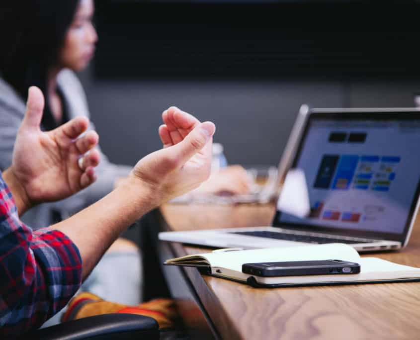 Person gesturing whilst discussing at a table with a laptop, notebook, and mobile in a meeting room.