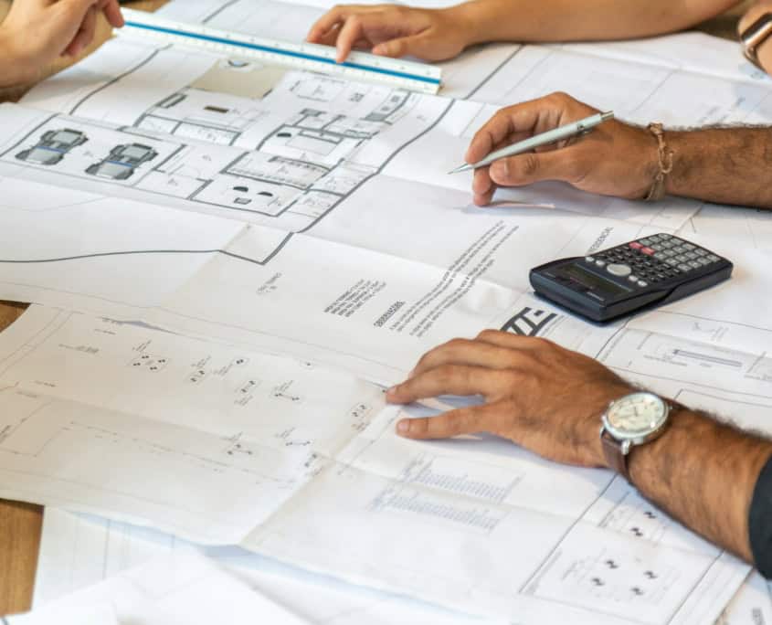 Two people review architectural blueprints on a table with a ruler, calculator, and documents.