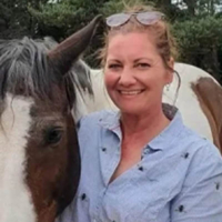 Woman smiling next to a brown and white horse outdoors, with trees in the background.