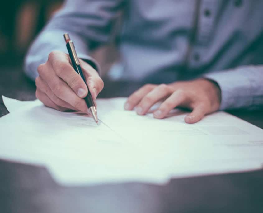 Person in a blue shirt writing on paper with a pen at a desk.
