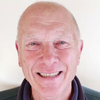Smiling older man with short, fair hair, wearing a dark collared shirt, against a plain background.